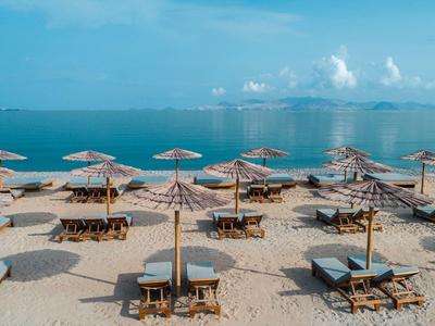 Strand mit mehreren Sonnenschirmen, Liegestühlen und ruhigem blauem Meer im Hintergrund.