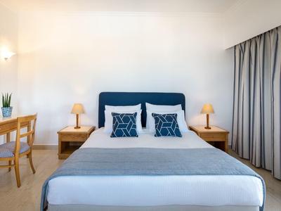 Hell eingerichtetes Schlafzimmer mit großem Bett, blauer Kopfstütze, Nachttischen und Vorhängen.