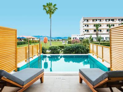 Schwimmbad mit blauer Wasserfläche, zwei Liegestühlen und Sicht auf Palmen und Gebäude im Hintergrund.