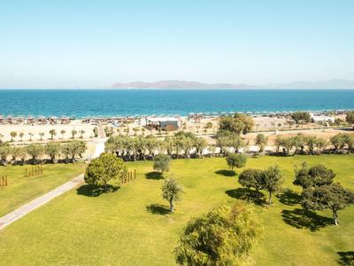 Grüner Park mit Bäumen vor einem Strand mit Sonnenschirmen und Meer im Hintergrund.