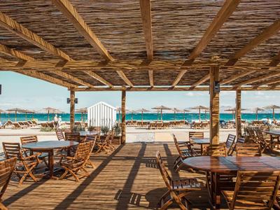 Offene Strandbar mit Holzstühlen und Tischen mit Blick auf Meer und Liegestühle bei Sonnenschein.