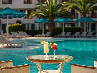 Zwei bunte Cocktails auf einem runden Tisch vor einem blauen Pool mit Liegestühlen und Palmen.