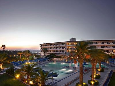 Hotel mit Außenpool, Palmen und beleuchteter Terrasse bei Sonnenuntergang.