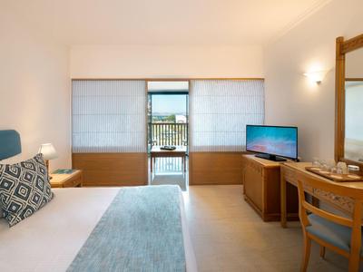 Helles Hotelzimmer mit Doppelbett, Schreibtisch, Stuhl und Blick auf einen Balkon mit Meerblick.