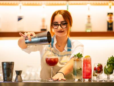 Barkeeperin gießt Getränk in Glas, bunte Cocktails und frische Zutaten im Hintergrund.