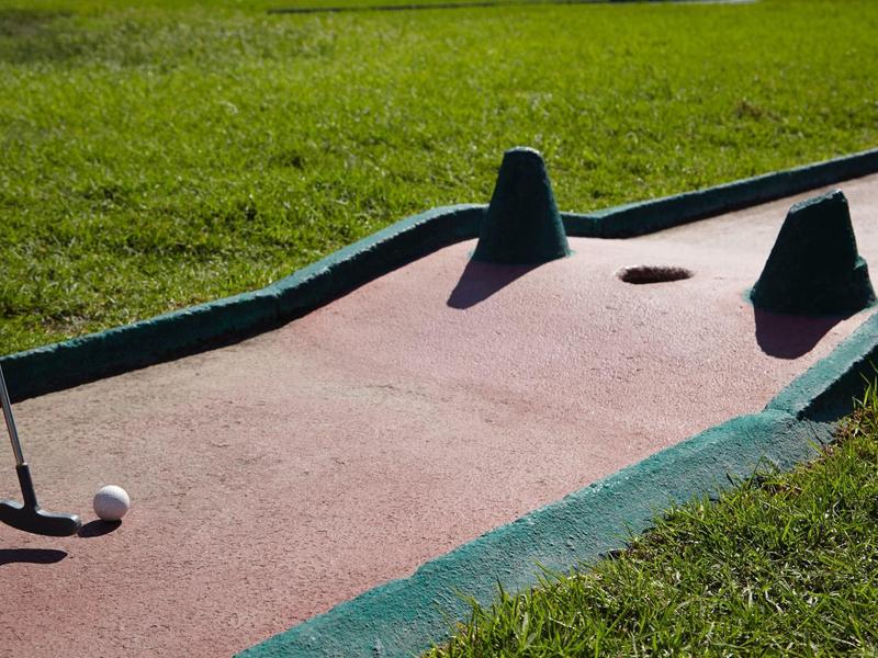 Minigolfbahn mit grünem Rand, zwei Hindernissen und einem Golfball auf rötlichem Boden.