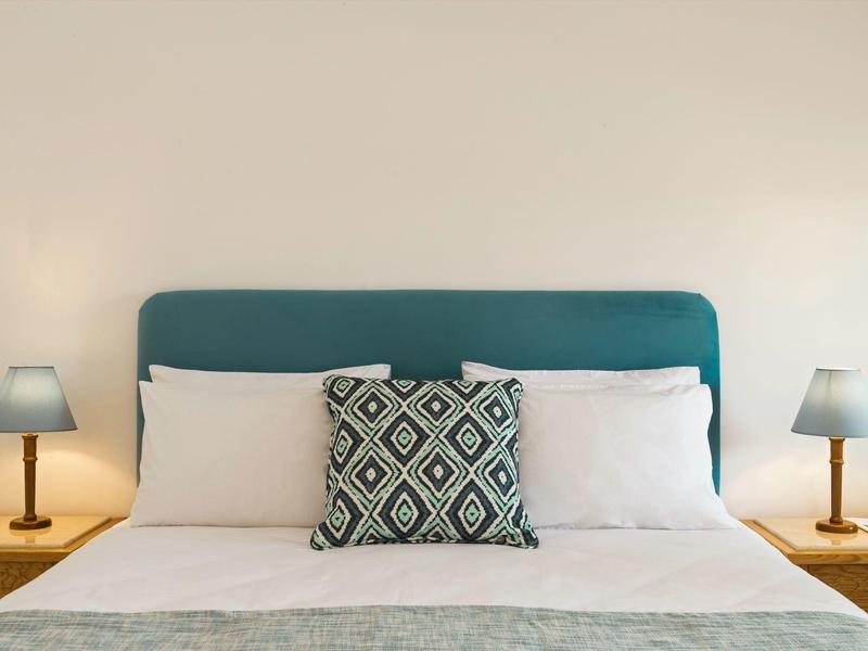 Bett mit türkisfarbenem Kopfteil, drei weißen Kissen und einem gemusterten Kissen, zwei Nachttischlampen.