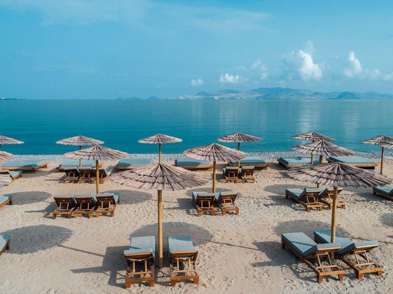 Strand mit mehreren Sonnenschirmen, Liegestühlen und ruhigem blauem Meer im Hintergrund.