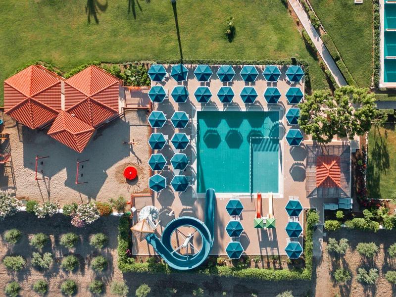 Vogelperspektive eines rechteckigen Pools mit blauen Liegestühlen, Schirmen und roten Dächern.