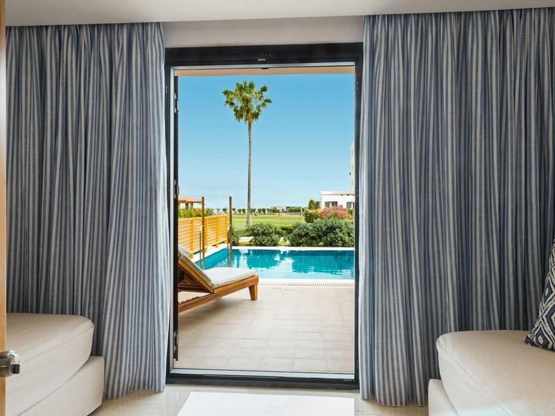 Blick durch offene Tür auf Pool mit Liegestuhl, Palmen und klarem Himmel, innen graue Vorhänge.