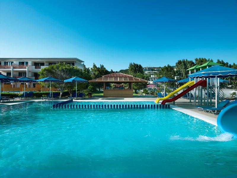 Großer blauer Pool mit Rutschen, Sonnenschirmen und niedrigem Gebäude im Hintergrund bei klarem Himmel.