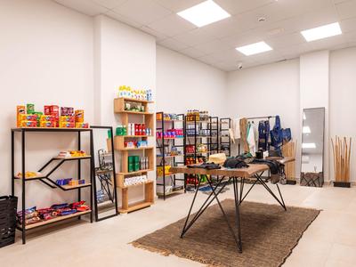 Jasno oświetlone pomieszczenie z półkami pełnymi kolorowych opakowań i stołem na środku na szarym dywanie.