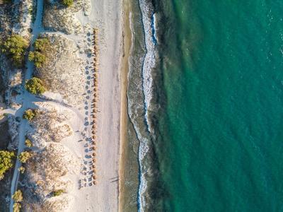 Luftaufnahme eines Strandes mit Liegestühlen und blauem Meer neben einem bewachsenen Dünenbereich.