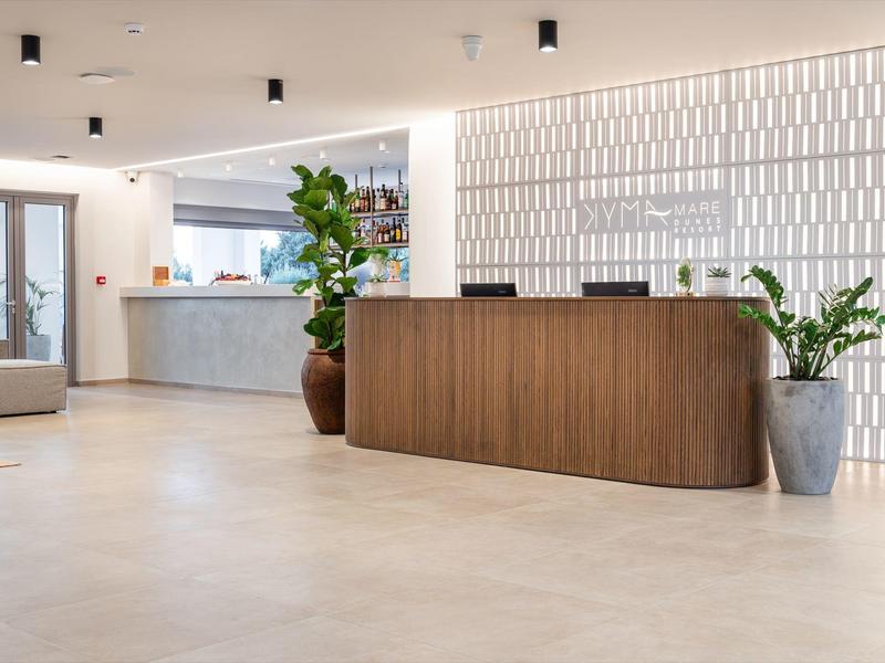 Nowoczesna recepcja hotelowa z drewnianym biurkiem i dużymi roślinami doniczkowymi w jasnym, minimalistycznym holu.