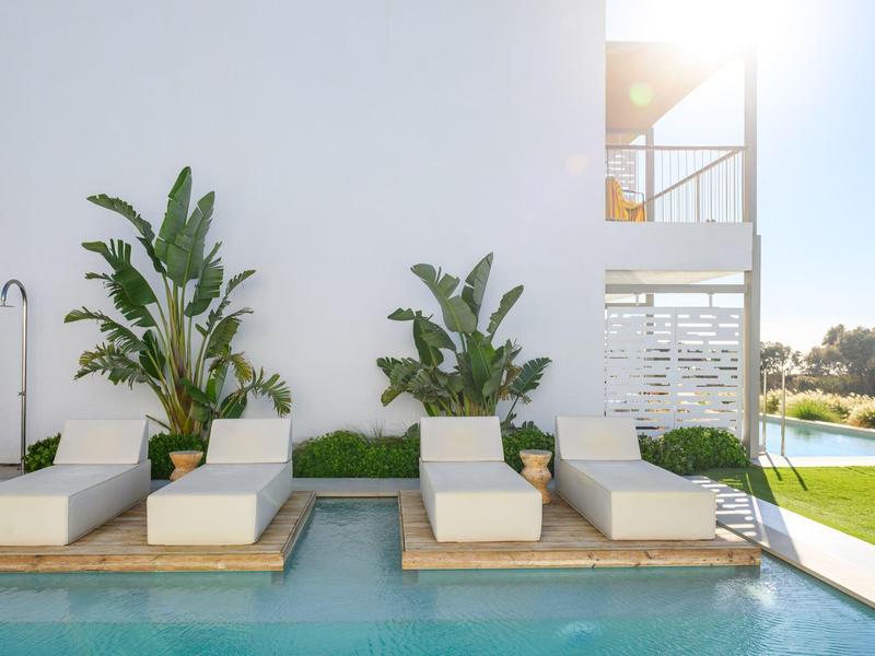 Nowoczesny obszar z basenem, białymi leżakami i dużymi roślinami doniczkowymi obok białego budynku.