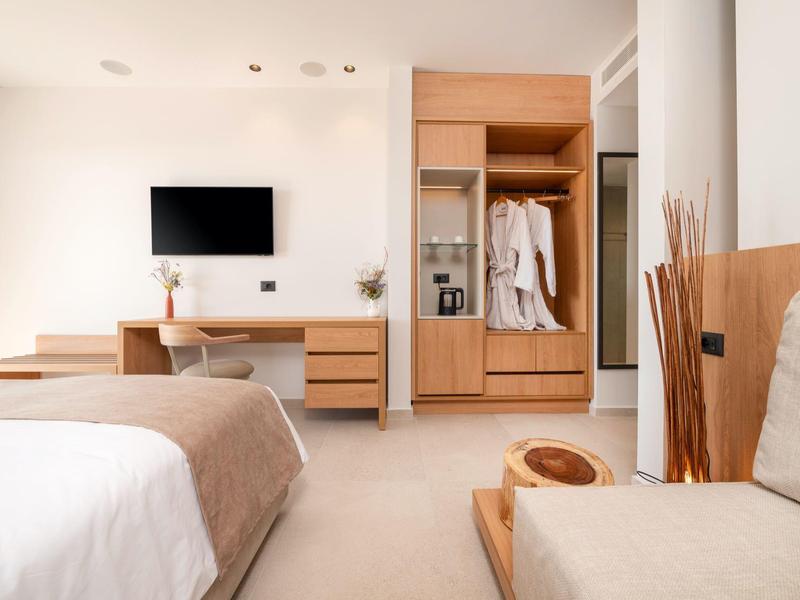 Nowoczesny pokój hotelowy z łóżkiem, telewizorem, biurkiem i otwartą szafą w neutralnych kolorach.