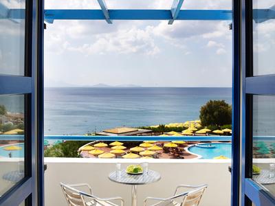 Balkon mit weißen Stühlen, Tisch, Meer und Pool mit gelben Sonnenschirmen im Hintergrund.