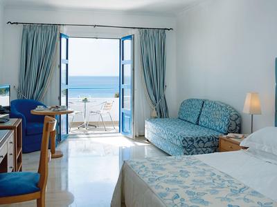 Hell eingerichtetes Schlafzimmer mit blauem Sofa, Bett, Stuhl und Blick auf das Meer durch offene Balkontür.