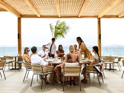 Menschen sitzen an einem Tisch im offenen Restaurant mit Blick aufs Meer unter Dach aus Holz.