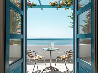 Blick durch offene blaue Türen auf Balkon mit Tisch, zwei Stühlen und Meer im Hintergrund.