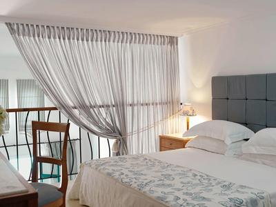 Moderní hotelový pokoj s manželskou postelí, šedým čelem a průsvitnou záclonou u okna.