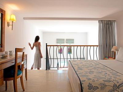 Žena v bílých šatech stojí na balkoně světlého, moderního hotelového pokoje s manželskou postelí.