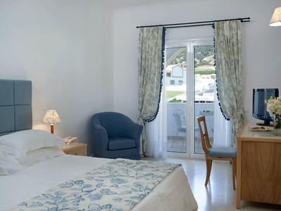 Helles Schlafzimmer mit blauem Sessel, Holzmöbeln und Balkon mit Blick auf weiße Häuser.
