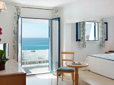 Helles Schlafzimmer mit Meerblick, offenem Balkon, kleinem Holztisch, Stühlen und weißen Möbeln.