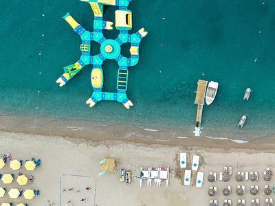 Luftaufnahme eines Strandes mit gelben Sonnenschirmen, Liegestühlen und einer großen blauen Wasserspielanlage.