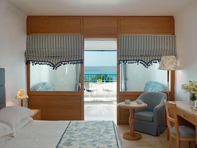 Helles Zimmer mit Doppelbett, blauem Sessel, Holzmöbeln und Blick auf Meer und Terrasse.