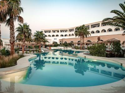 Hotel mit mehreren Stockwerken, Pool, Palmen und Liegestühlen in tropischer Umgebung bei Sonnenuntergang.