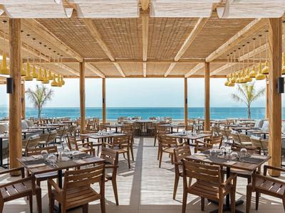 Offenes Restaurant mit Holzmöbeln und Blick auf das Meer unter einem Holzpergola-Dach.