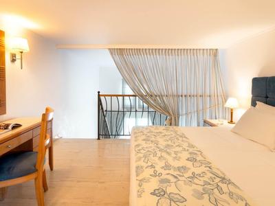 Hell eingerichtetes Hotelzimmer mit großem Bett, Schreibtisch und Balkon mit Vorhängen.