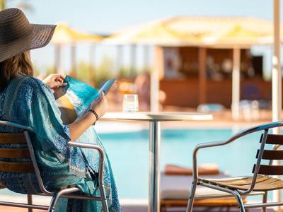 Frau im Hut sitzt entspannt auf Terrasse und liest Buch mit Pool und Sonnenschirmen im Hintergrund.