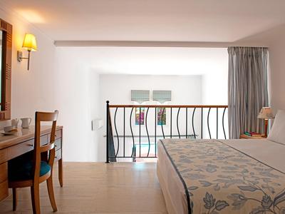 Helles Hotelzimmer mit Bett, Holzschreibtisch, Stuhl und Balkon mit Geländer und Vorhängen.