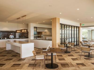 Moderne Hotel-Lounge mit Sitzbereichen, Tischen, Bar und offenen Küchenbereich in warmen Farben.