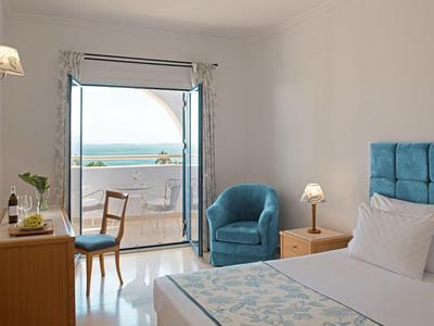 Helles Hotelzimmer mit Doppelbett, blauem Sessel und Balkon mit Meeresblick.