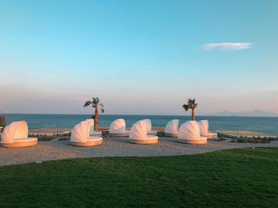 Strand mit grasbewachsenem Bereich, fünf weißen Lounge-Sitzen und Palmen unter klarem Himmel.