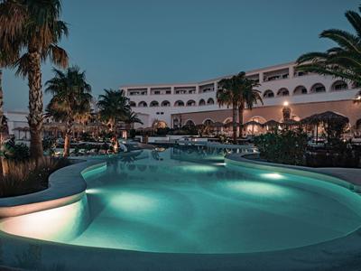Ein beleuchteter Pool vor einem mehrstöckigen Hotelgebäude bei Abenddämmerung mit Palmen im Vordergrund.