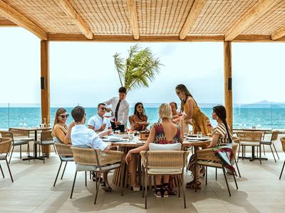 Menschen genießen Mahlzeit auf offener Terrasse mit Meerblick und Palmen.