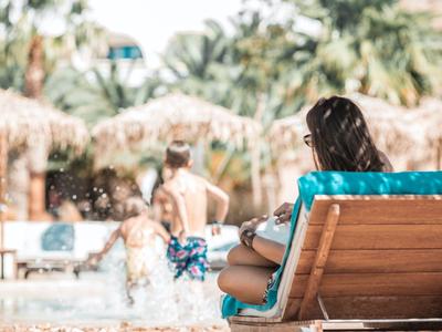 Frau sitzt auf einer Liege am Pool in einem tropischen Urlaubsort.