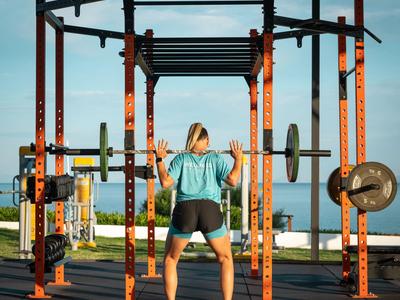 Frau trainiert mit Langhantel an rotem Outdoor-Fitnessgerät mit Meerblick.