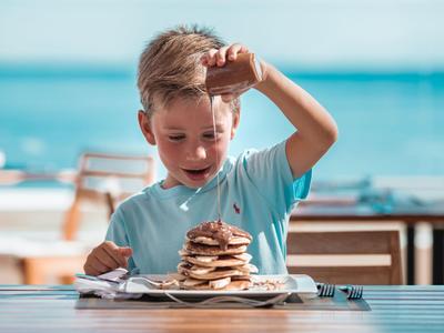 Ein Junge sitzt am Tisch vor einem großen Stapel Pfannkuchen mit Blick aufs Meer.