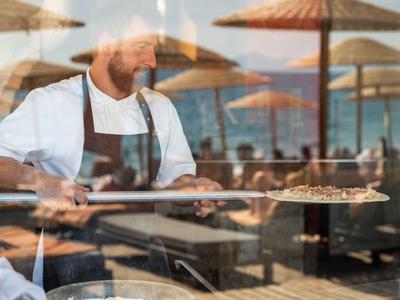 Koch serviert frisch zubereitete Pizza in einem Restaurant mit Meerblick und Sonnenschirmen.