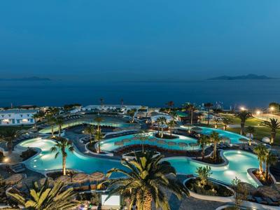 Großer Resort-Pool mit beleuchteten Wasserflächen und Palmen am Meer bei Nacht.