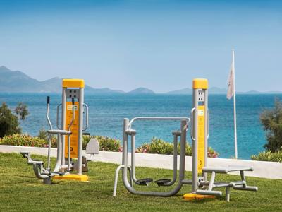 Outdoor-Fitnessgeräte stehen auf einem Rasen mit Blick auf Meer und Berge.