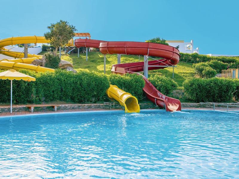 Bazén se žlutými a červenými tobogány na pozadí zelených kopců a modré oblohy.