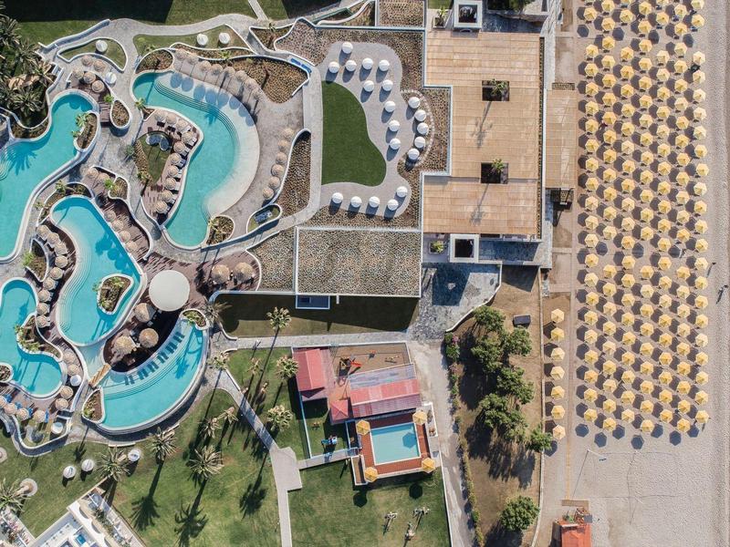 Letecký pohled na resort s klikatým bazénem, lehátky a písečnou pláží u moře.