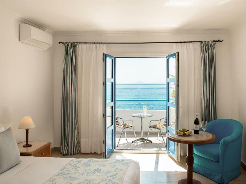 Světý hotelový pokoj s výhledem na moře, modré křeslo, malý stůl a balkon s výhledem na oceán.