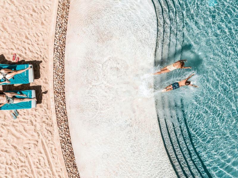 Lidé odpočívající na lehátkách a plavající v zakřiveném bazénu vedle písečné pláže